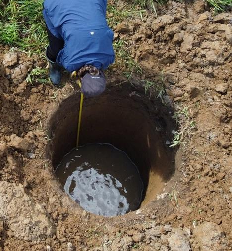 An archaeologist measures a flooded excavation pit with water visible at the bottom