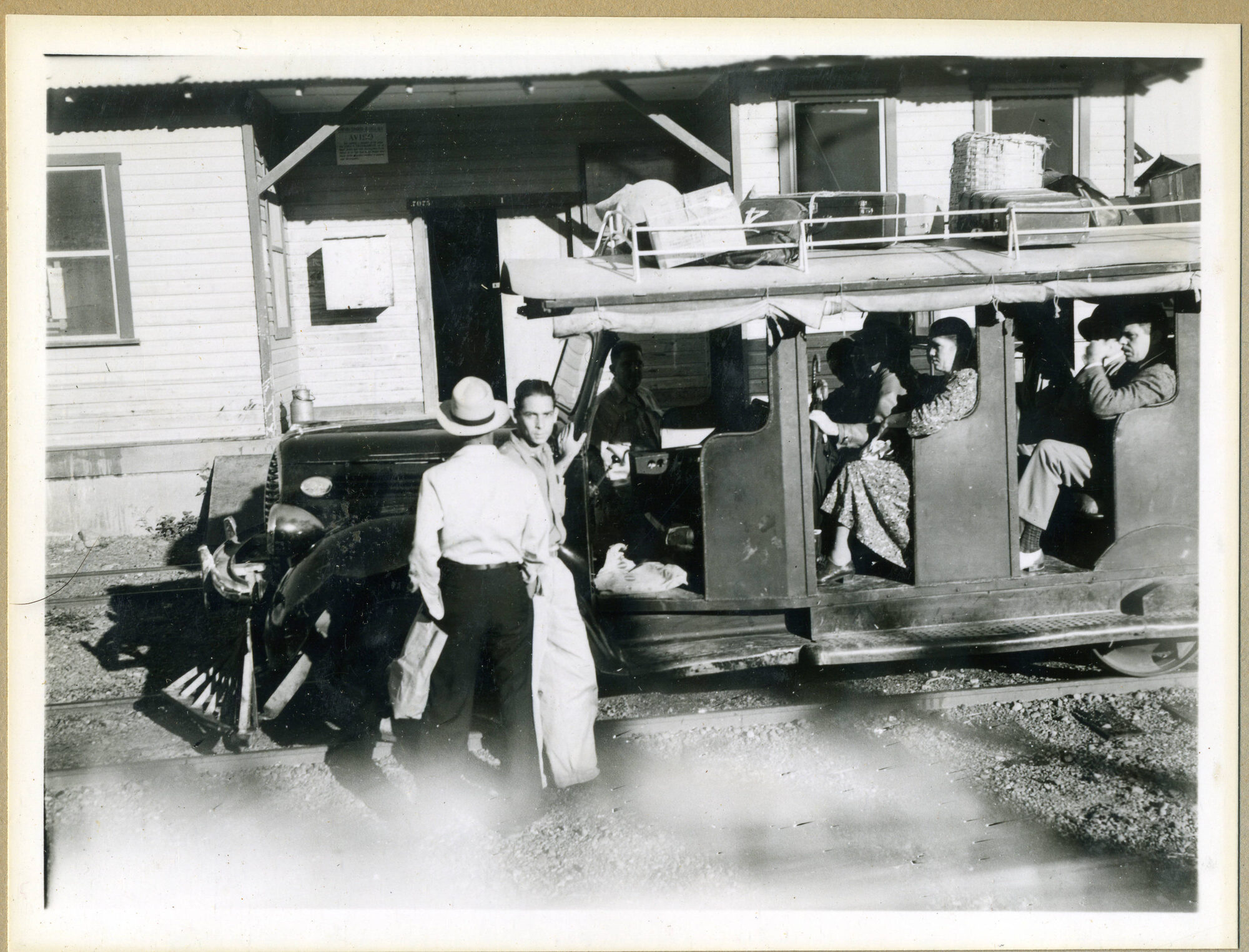Black and white photograph of a modified Dodge sedan on railroad tracks, carrying passengers and cargo at a company station